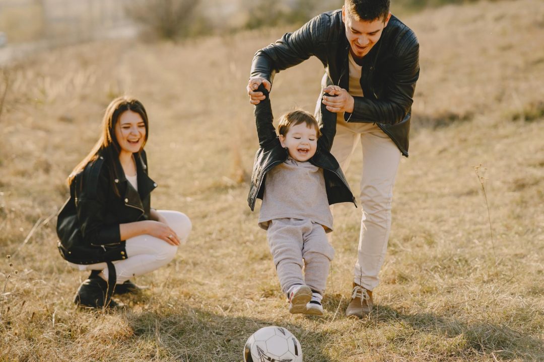 Father and son are playing football, mother is watching.