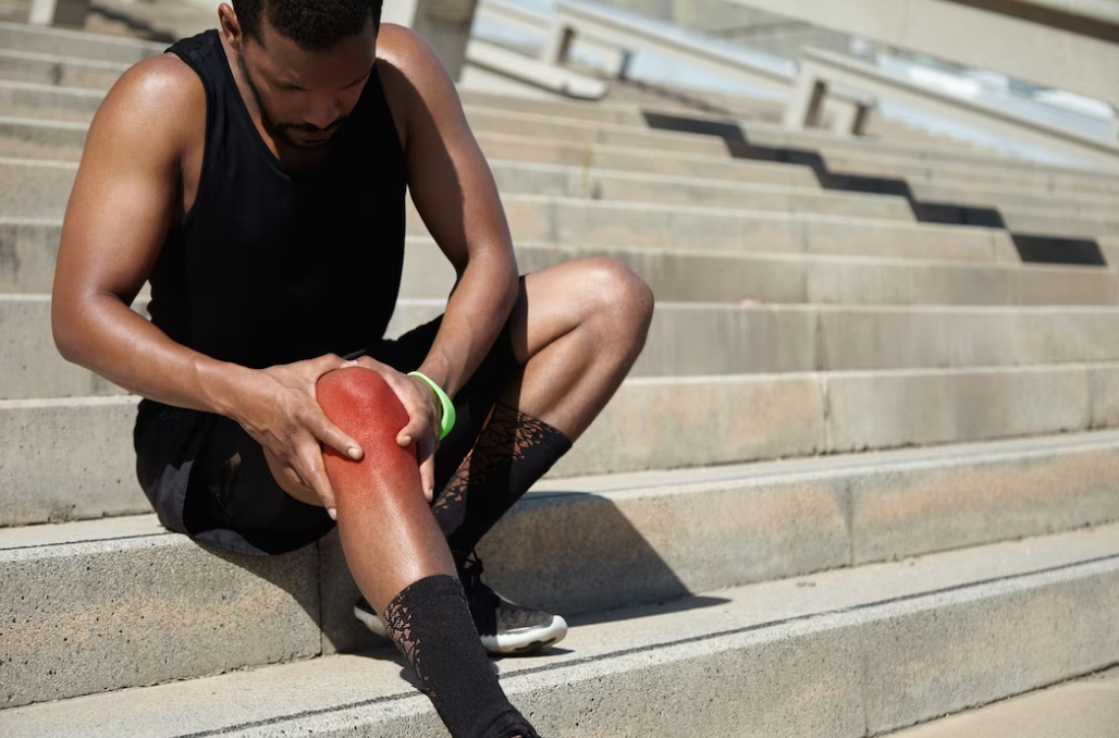 man in a black t-shirt and shorts sitting outside on the stairs and holding his knee