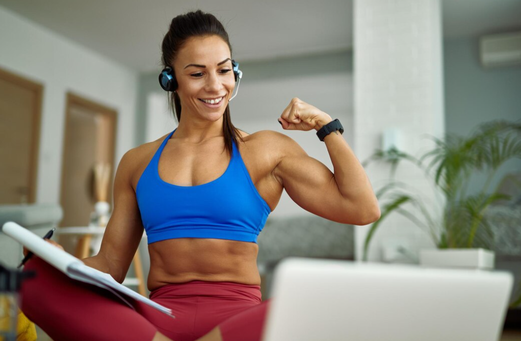 woman in a blue t-shirt and red leggings flexing her bicep while making video call over a laptop at home