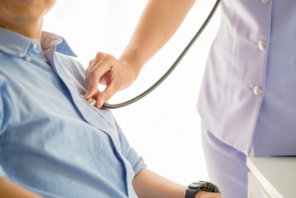 man in blue shirt sitting in a chair, woman with stethoscope listening to his heartbeat